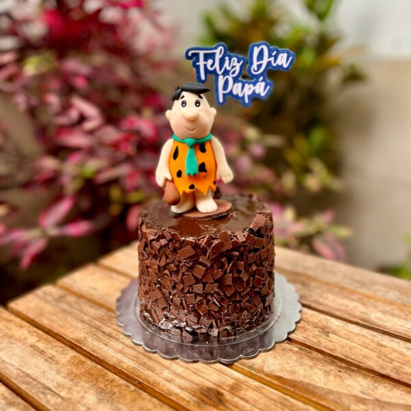 Torta decorada con trocitos de chocolate y Pedro Picapiedra en fondant sobre la parte superior, acompañada de un topper que dice “Feliz Día, Papá”.