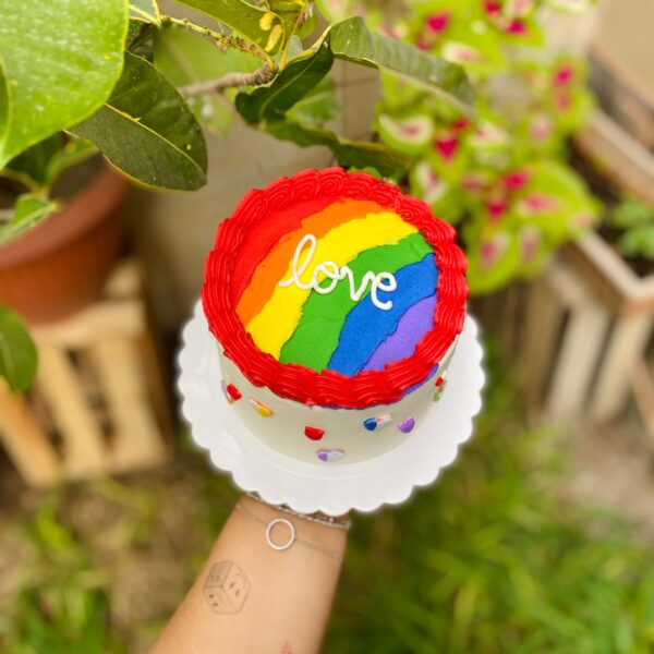 Torta temática Pride decorada con buttercream blanco, rosetones rojos y corazones con los colores de la bandera del Pride. En la parte superior lleva la palabra "Love" sobre una franja multicolor.