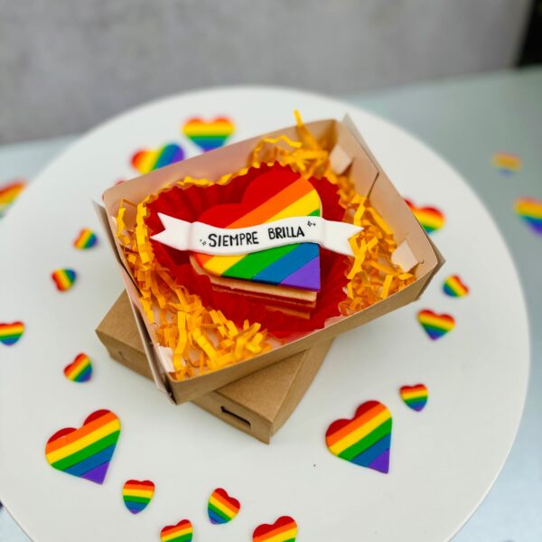 Alfajor en forma de corazón decorado con fondant en los colores de la bandera Pride y un banner con la frase “Siempre Brilla”.