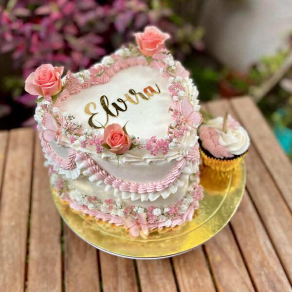 Torta en forma de corazón decorada con rosetones de buttercream en distintos tonos, flores naturales, mariposas de cartulina y un cupcake de la misma temática al costado.