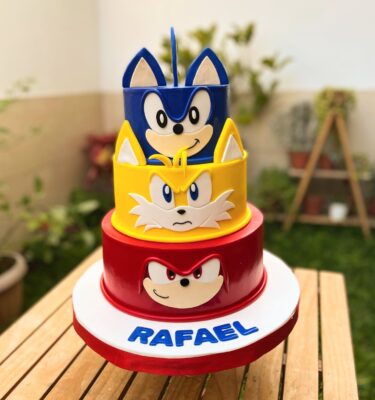 Torta de tres pisos en fondant, cada uno representando a un personaje: piso inferior rojo con la cara de Knuckles, piso intermedio amarillo con la cara de Tails y piso superior azul con el rostro de Sonic.