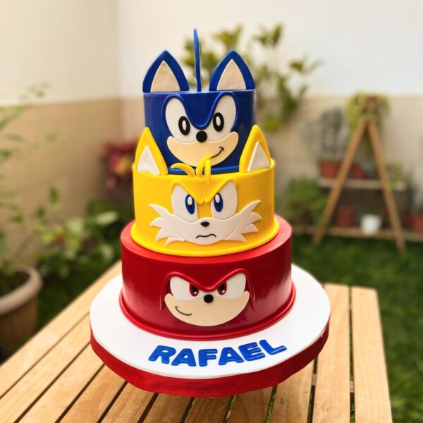 Torta de tres pisos en fondant, cada uno representando a un personaje: piso inferior rojo con la cara de Knuckles, piso intermedio amarillo con la cara de Tails y piso superior azul con el rostro de Sonic.