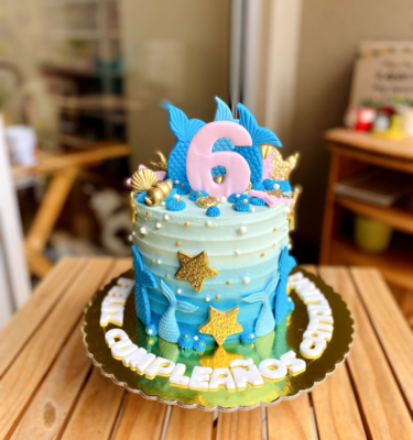 Torta de un piso bañada en buttercream, decorada con colas de sirena, conchitas y estrellas de mar en fondant, en tonos marinos y detalles modelados a mano.