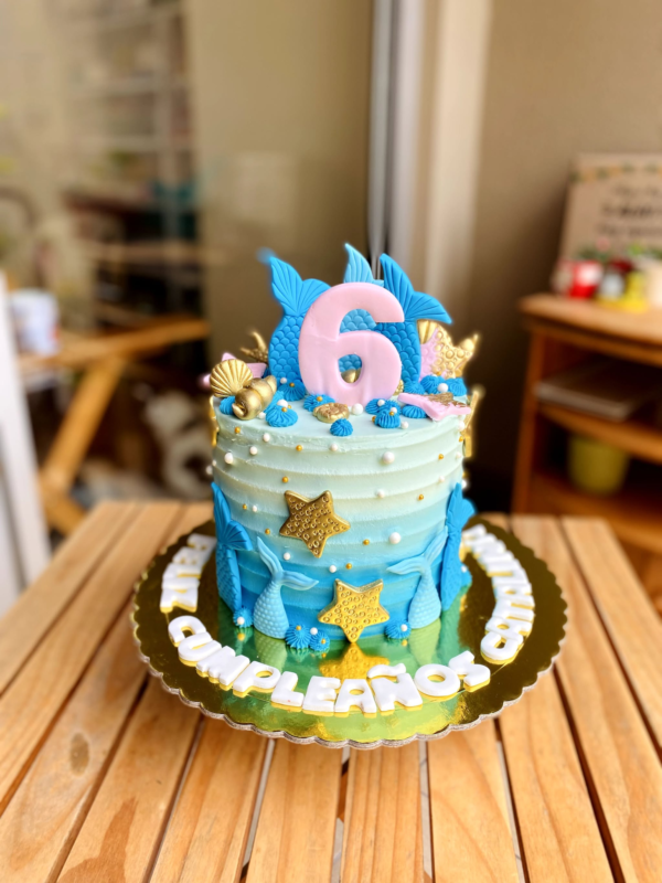 Torta de un piso bañada en buttercream, decorada con colas de sirena, conchitas y estrellas de mar en fondant, en tonos marinos y detalles modelados a mano.