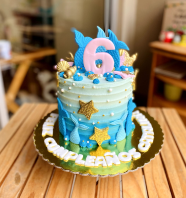 Torta de un piso bañada en buttercream, decorada con colas de sirena, conchitas y estrellas de mar en fondant, en tonos marinos y detalles modelados a mano.