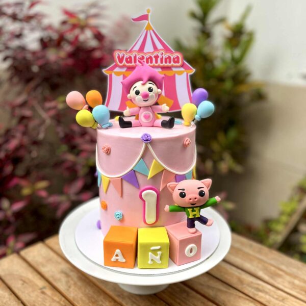 Torta de un piso en tonos rosados con temática de Plim Plim. Lleva una figura de Plim Plim rosado en la parte superior, el cerdito en la base, globos decorativos, banderines y pequeñas flores.
