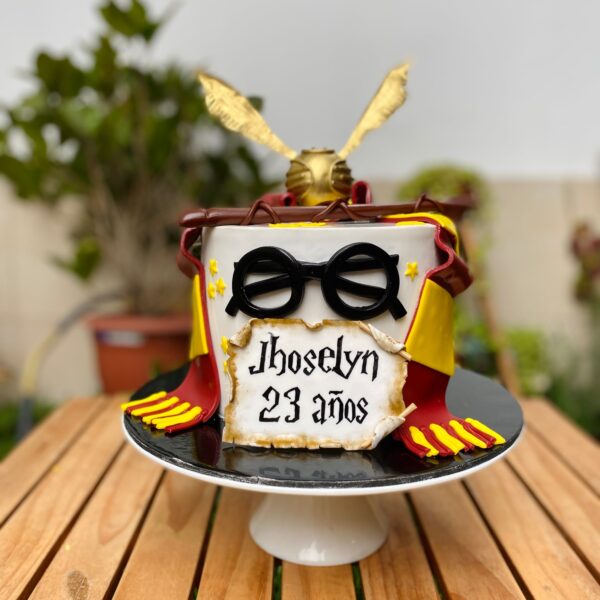 Torta temática de Harry Potter con bufanda de Gryffindor, varita mágica, lentes, Snitch dorada y pergamino personalizado en fondant.