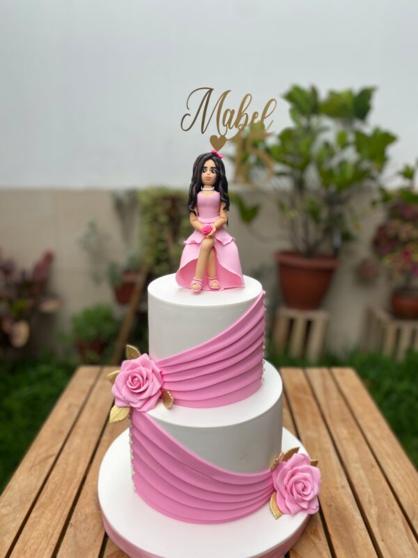 Torta de dos pisos forrada en fondant, decorada con rosas de azúcar, drapeado elegante y una muñeca en 3D de fondant de la cumpleañera, acompañada de topper dorado personalizado.