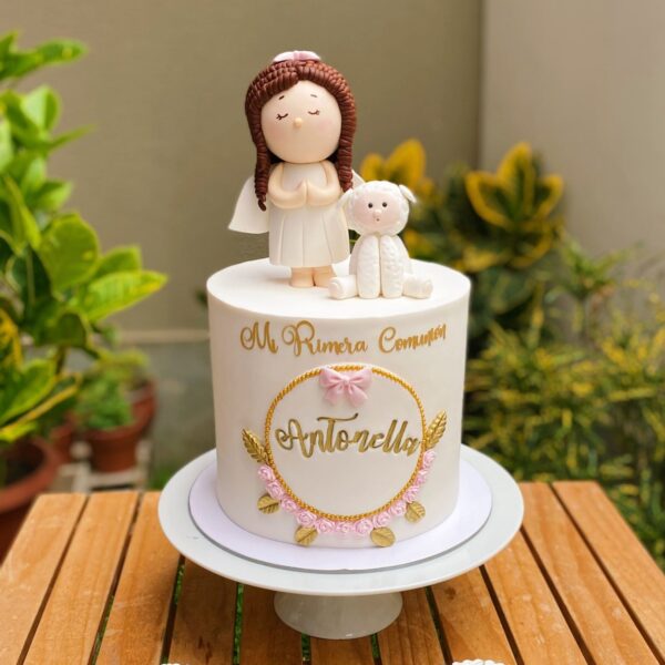 Torta de un piso para Primera Comunión, con una angelita y ovejita en 3D de azúcar, marco de fondant con flores, detalles en dorado y frase “Mi Primera Comunión”.