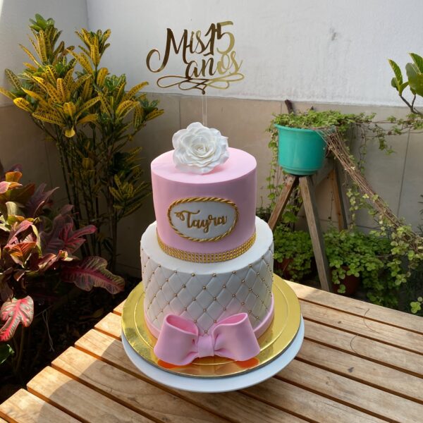 Torta de dos pisos forrada en fondant, con base en diseño capitoné, un moño decorativo alrededor, nombre personalizado en el segundo nivel y una rosa de azúcar en 3D como topper.