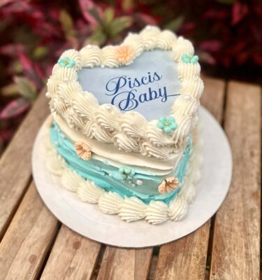 Burncake de Piscis decorado con buttercream en colores tropicales, ondas que simulan olas del mar, flores en fondant y perlas comestibles, con imagen personalizada en papel de arroz que se quema para revelar un mensaje sorpresa.