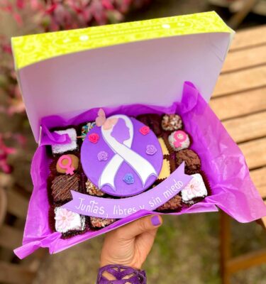 Mix box con 20 postres variados (brownies, alfajores y trufas) con decoración central en fondant del Día de la Mujer con lazo morado y silueta de rostro femenino, acompañado de un banner que dice “Juntas, libres y sin miedo”.
